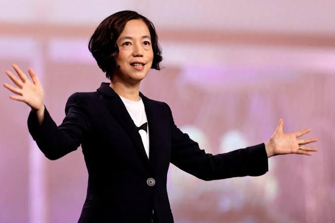 A speaker in a dark blazer with arms outstretched on a conference stage against a pink backdrop