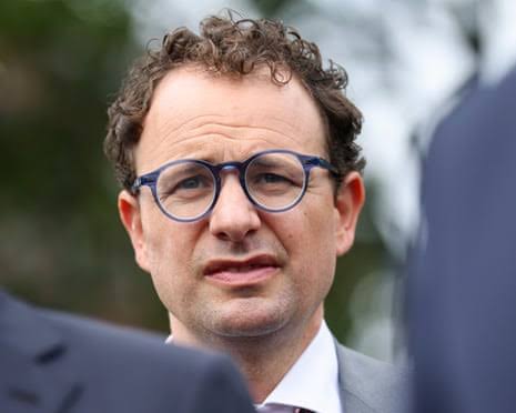 A man in round blue-rimmed glasses and a grey suit listens during an outdoor press briefing