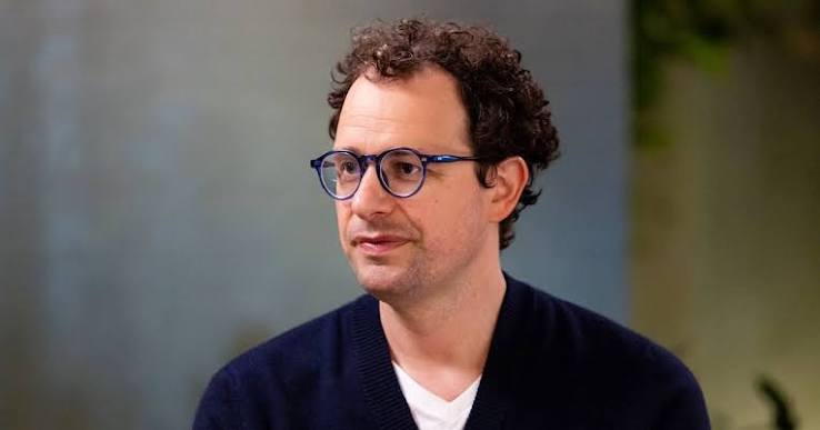 A man with curly hair and round blue-framed glasses in a navy sweater, photographed in an interview setting