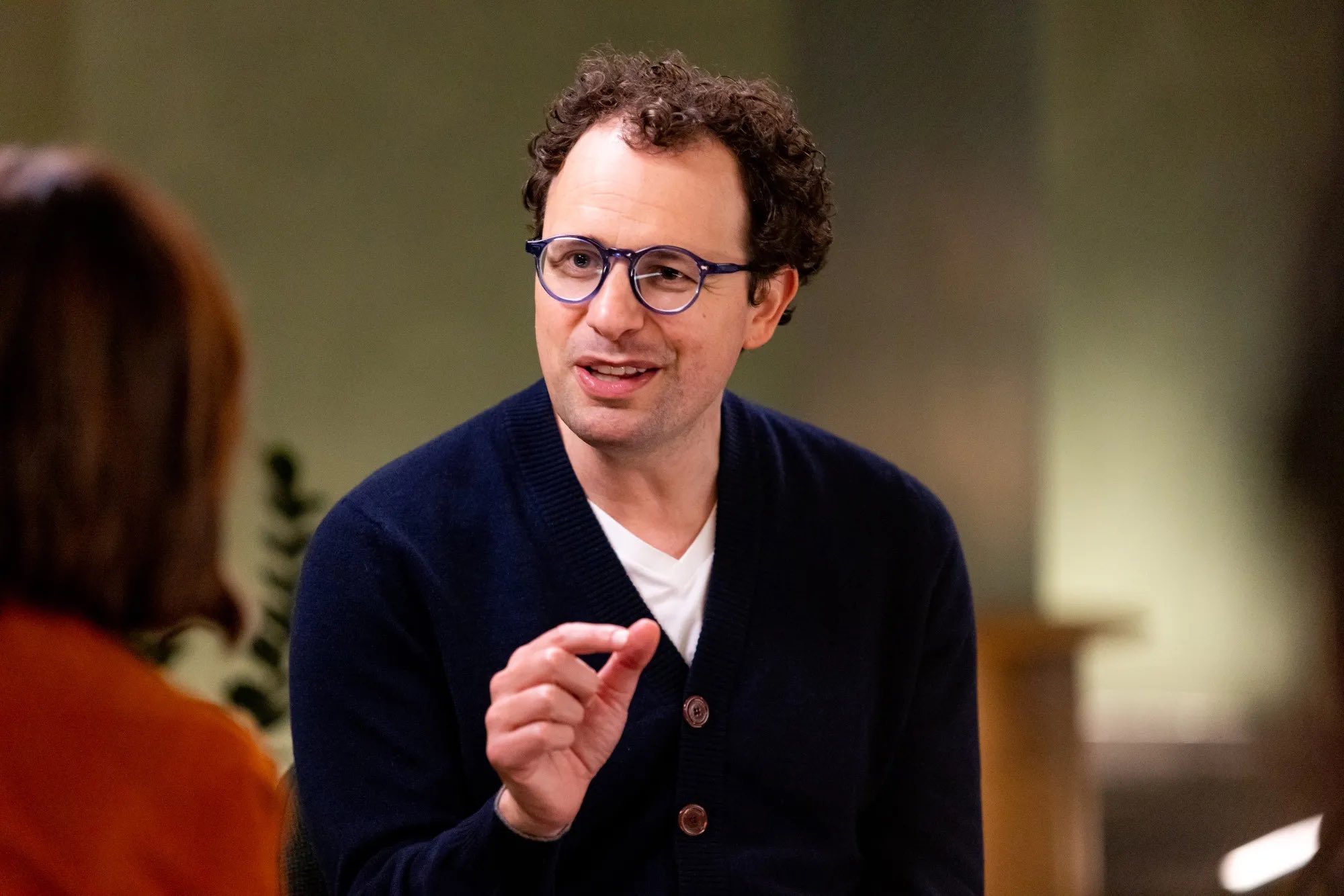 A man in glasses and a navy cardigan speaks during a seated interview