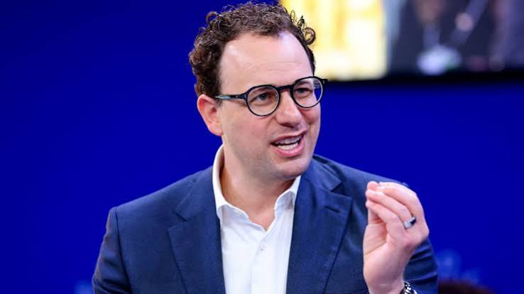 A man in glasses and a navy blazer speaking and gesturing on stage against a blue backdrop