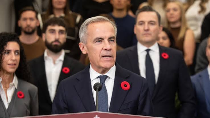 Prime Minister Mark Carney speaks at a podium, wearing a remembrance poppy, with people standing behind him