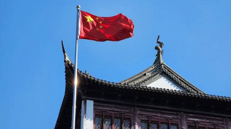 The national flag of China flying above a traditional Chinese building with a curved tiled roof against a clear blue sky