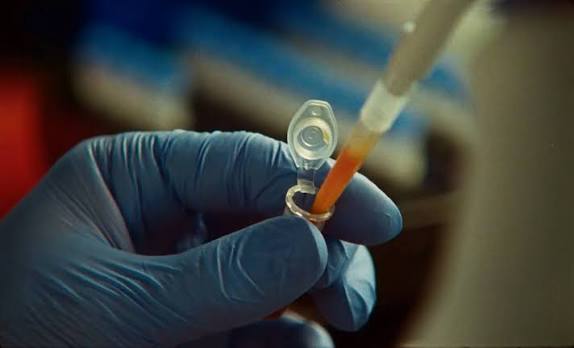 A scientist in a blue nitrile glove pipettes an orange liquid into a small laboratory tube
