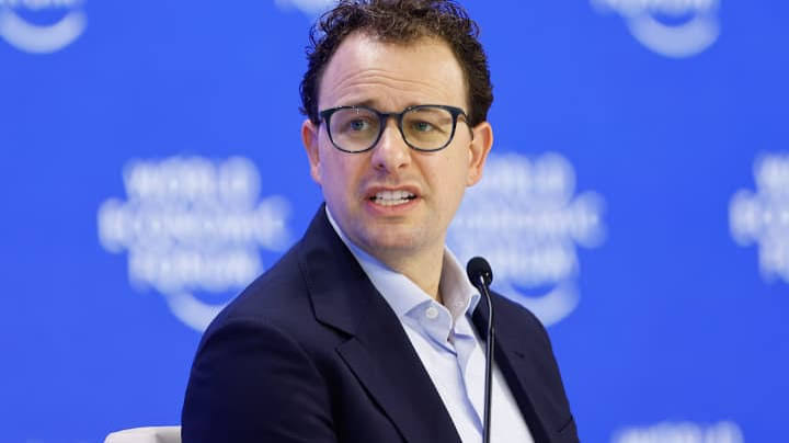 A speaker in glasses and a dark jacket addresses a World Economic Forum panel in front of a blue backdrop