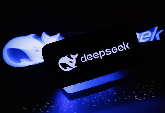 DeepSeek whale logo and wordmark glowing on a phone screen held in front of a laptop, with a blurred DeepSeek logo in the background