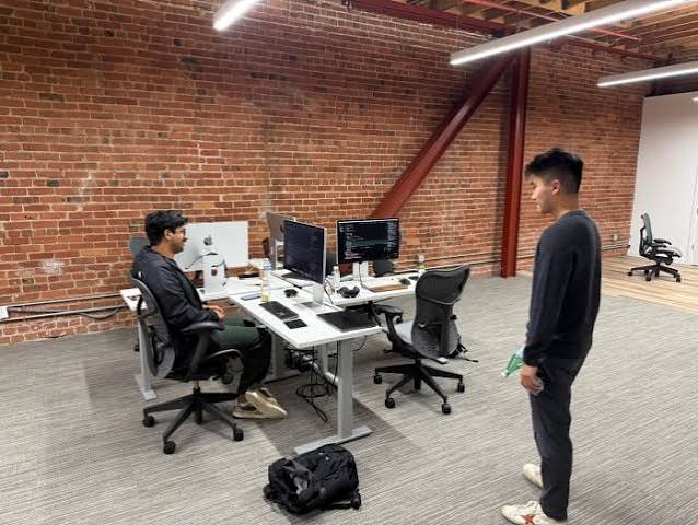 Two young founders in a brick walled startup office, one seated at a desk with code on the monitors and another standing nearby