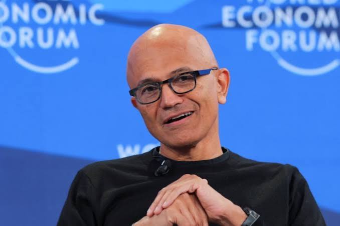 Microsoft CEO Satya Nadella in glasses and a black sweater speaking on stage in front of a blue World Economic Forum backdrop