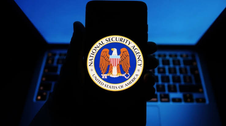 A hand holding a smartphone showing the National Security Agency seal in front of a glowing laptop keyboard