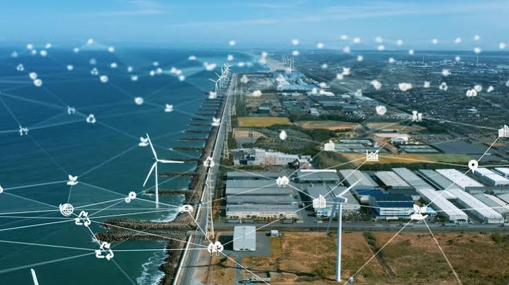 An aerial view of a coastal industrial park with wind turbines and warehouses, overlaid with a glowing network of connected energy icons