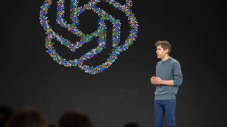 A speaker in a grey sweater and jeans on stage beside a large OpenAI logo made of multicolored dots