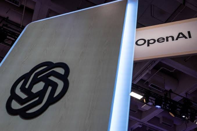 The OpenAI logo on an illuminated display pillar beside an overhead OpenAI sign at a technology event