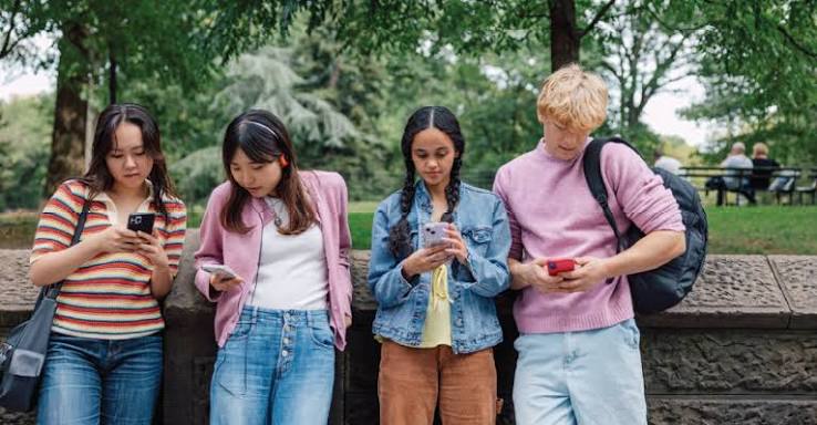 Four teenagers stand against a stone wall in a park, each looking down at a smartphone