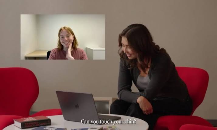 A woman on a red chair smiles at a laptop during a live video call with a Tavus Griffin AI persona shown as a young woman touching her chin, from Tavus's Griffin launch video