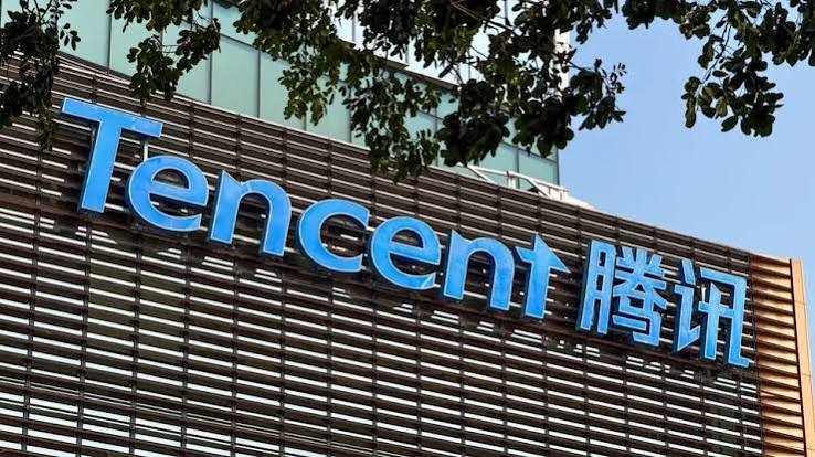 The blue Tencent logo and its Chinese name on the slatted facade of a Tencent office building, framed by tree branches