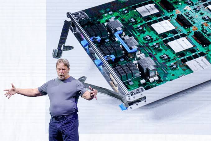 A presenter speaks on stage in front of a large projected image of an AI server board