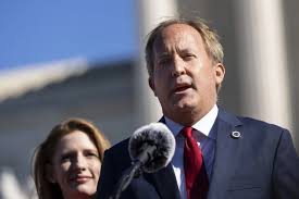 Texas Attorney General Ken Paxton speaking into a microphone at an outdoor press event, with a woman standing behind him