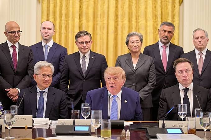 Technology executives and officials seated and standing around a meeting table in a gold-curtained White House room, with name placards for Nvidia's Jensen Huang and Speaker Mike Johnson