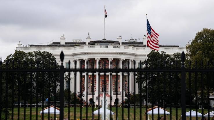 The White House in Washington behind its black iron fence under a grey sky, with the US flag flying on the South Lawn