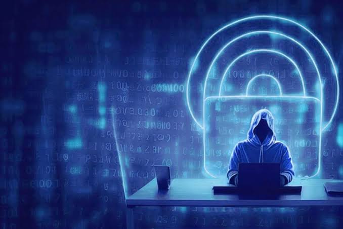 A hooded figure works on a laptop in front of a glowing blue padlock and streams of code